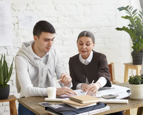 foto-sincera-dos-companeros-trabajo-que-intercambian-ideas-oficina-ingeniera-jefa-mediana-edad-que-demuestra-su-punto-vista-su-joven-colega-moreno-senalando-plano-frente-ella-escritorio foto-sincera-dos-companeros-trabajo-que-intercambian-ideas-oficina-ingeniera-jefa-mediana-edad-que-demuestra-su-punto-vista-su-joven-colega-moreno-senalando-plano-frente-ella-escritorio